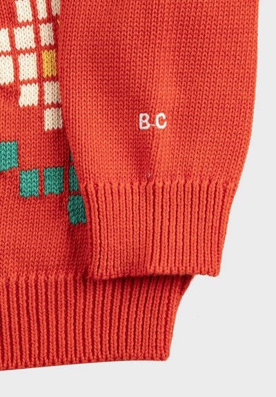 Primer plano de la manga de un suéter de punto naranja con puño acanalado y letras bordadas en blanco "B_C" cerca de la muñeca.