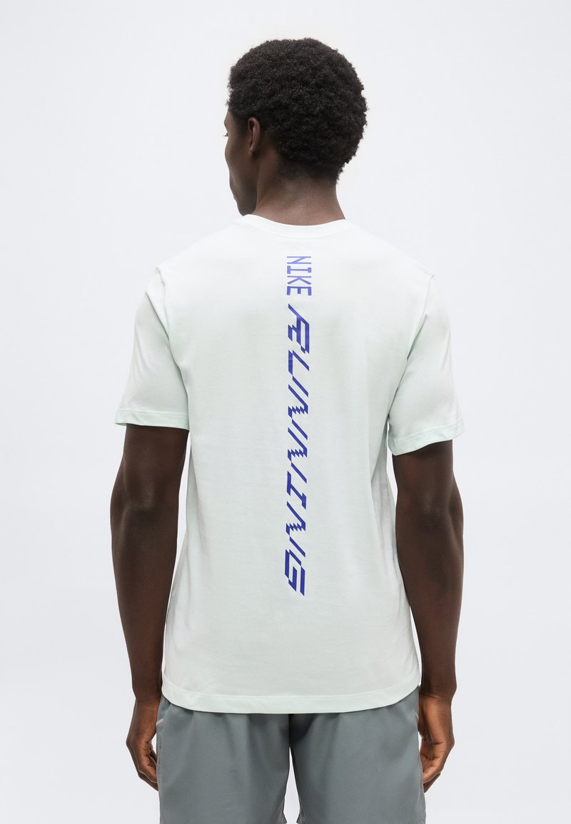 Άνδρας που φοράει λευκό αθλητικό T-shirt της Nike με μπλε κάθετη επιγραφή "NIKE RUNNING" στην πλάτη και γκρι σορτς, στέκεται μπροστά σε απλό φόντο.