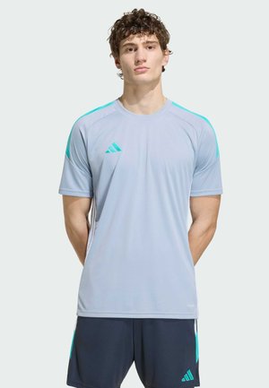 Giovane uomo in piedi con le braccia dietro la schiena, indossa una maglietta sportiva Adidas grigio chiaro e pantaloncini scuri con accenti turchesi.