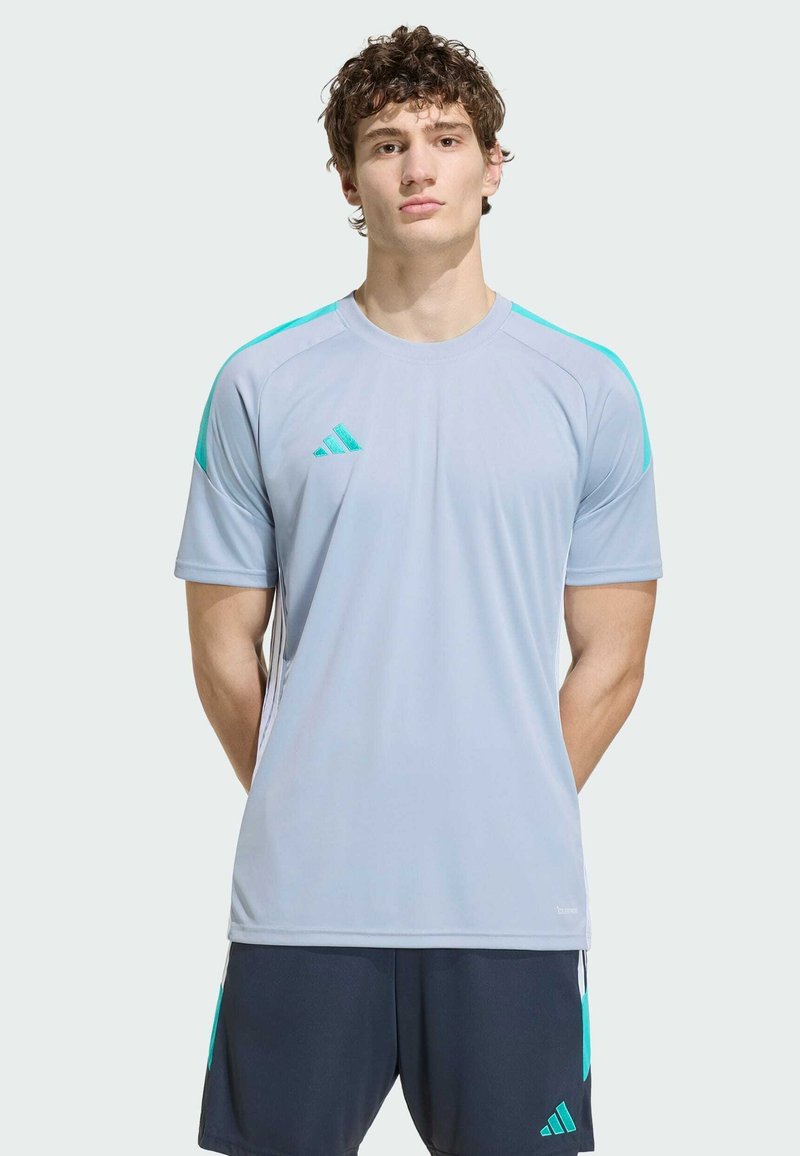 Jeune homme debout, les bras derrière le dos, portant un maillot de sport Adidas gris clair et un short foncé avec des accents turquoise.
