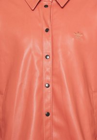 Giacca in pelle corallo con chiusura frontale a bottoni, bottoni neri e un discreto logo adidas dorato vicino al colletto. Texture liscia, design strutturato.