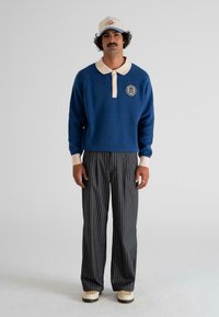 Man wearing blue sweater with beige collar, gray pinstripe pants, beige shoes, and a beige cap with text, standing against plain light background.