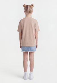 Bērns ar gaišiem matiem divos cukurvītos un bizītēs, tērpies bēša krāsas T-kreklā, džinsu svārkos, baltās zeķēs un baltos apavos, stāv ar muguru facing.