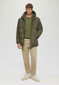 Puffjacke in Olivgrün mit roten Reißverschlussakzenten, kombiniert mit einem grünen Pullover und beigen Hosen. Weiße und beigen Sneakers runden den Look ab.