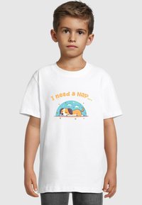 Hvid bomulds t-shirt med en tegneseriehund, der sover under en blå himmel, med teksten "Jeg har brug for en lur..." i orange ovenfor. Korte ærmer.