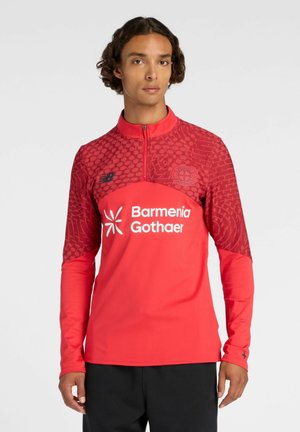Jeune homme portant un haut de sport rouge à manches longues avec le logo Barmenia Gothaer et un pantalon noir, debout devant un fond gris uni.