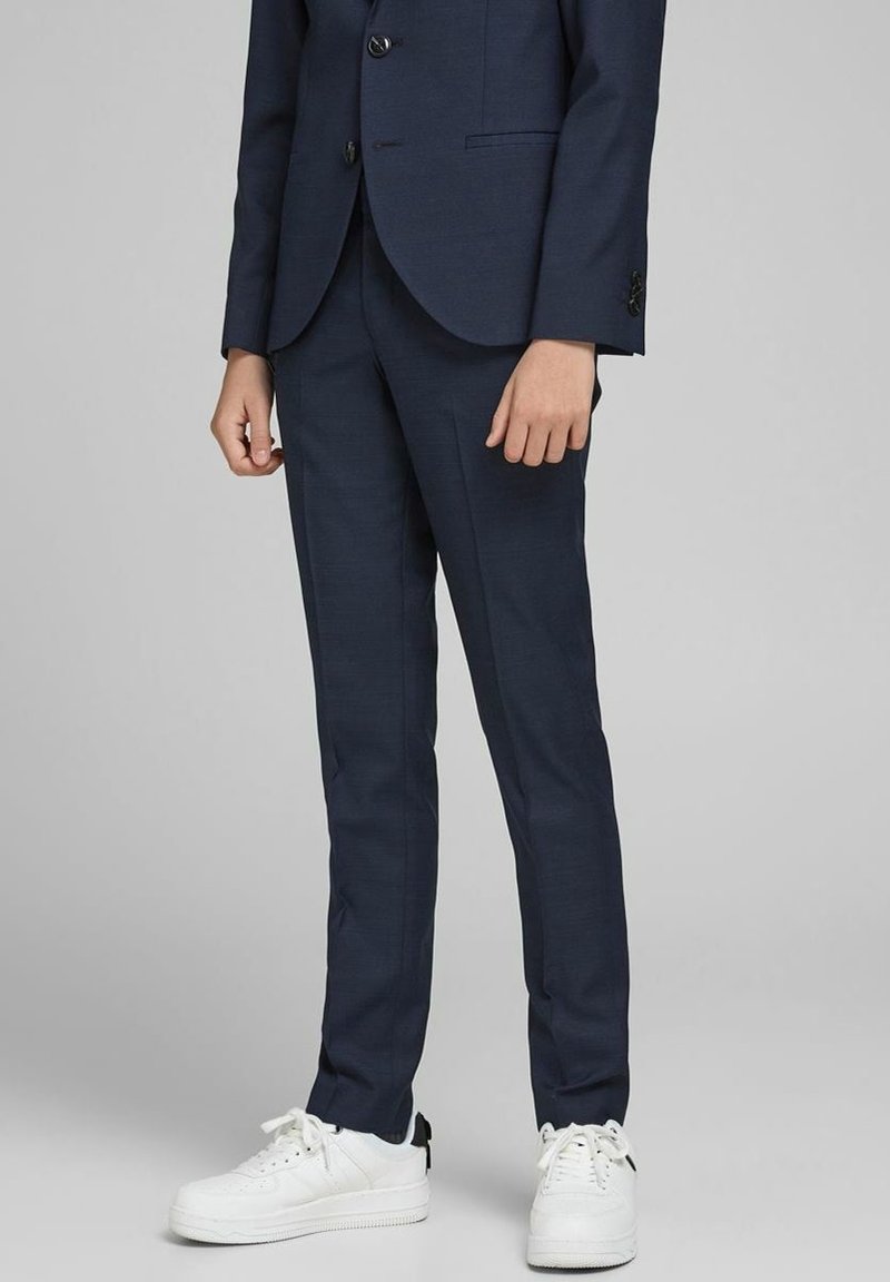 Tmavě modré kalhoty na oblek s úzkým střihem, texturovaná látka, spárované s bílými sportovními botami. Přední kapsy a jemné šití jako detaily.