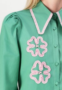 Camisa verde con un collar de volantes rosa y dos apliques florales rosas con acentos de perlas. Mangas largas con hombros abullonados.