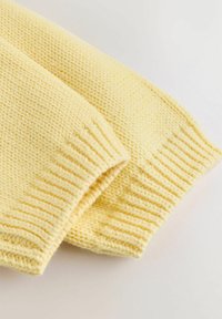 Rumena pletena jopica z rebrastimi manšetami, ki ima teksturiran vzorec in mehak material. Oblikovanje je preprosto in sproščeno.