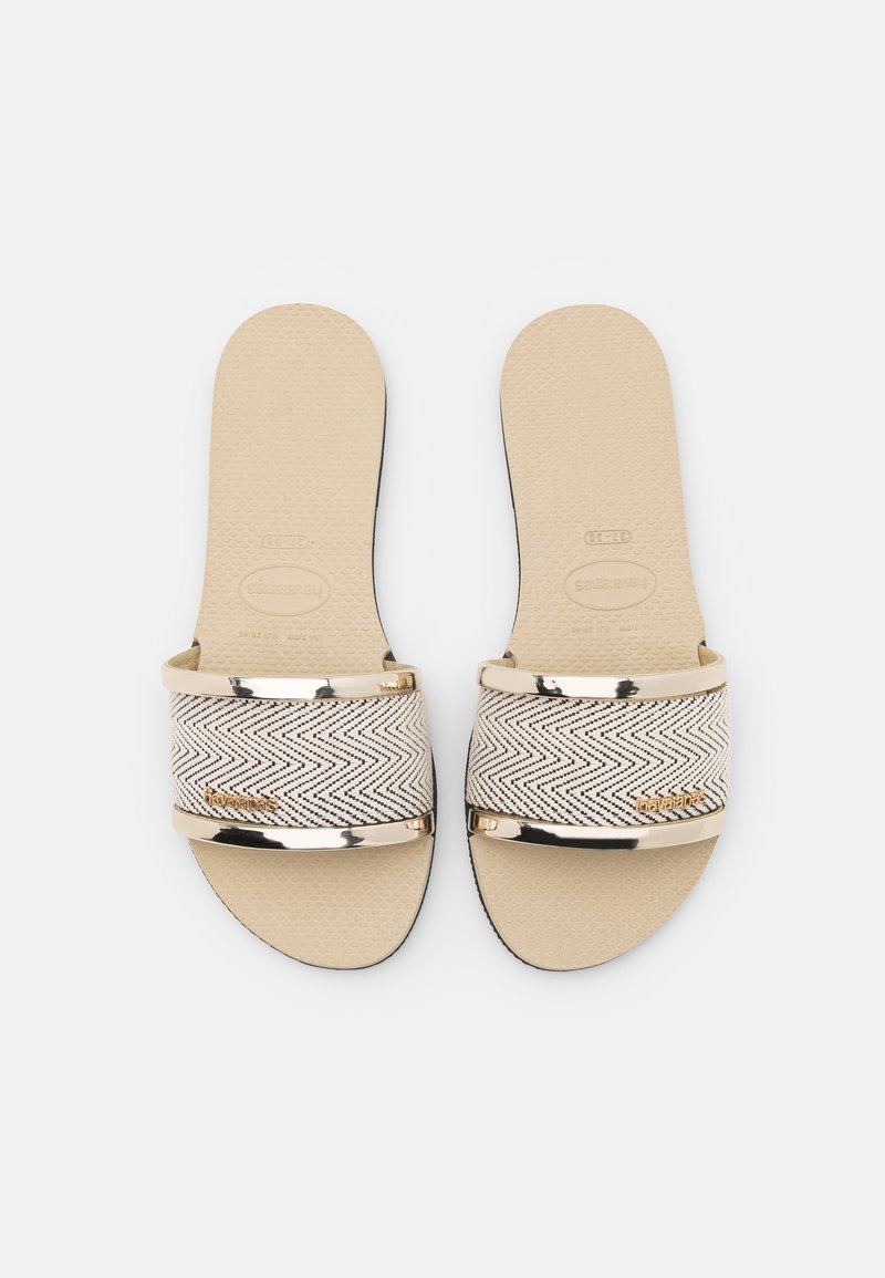 Paire de sandales plates beiges avec une bride en tissu noir à motif zigzag et une bordure dorée, vues de dessus sur fond blanc.