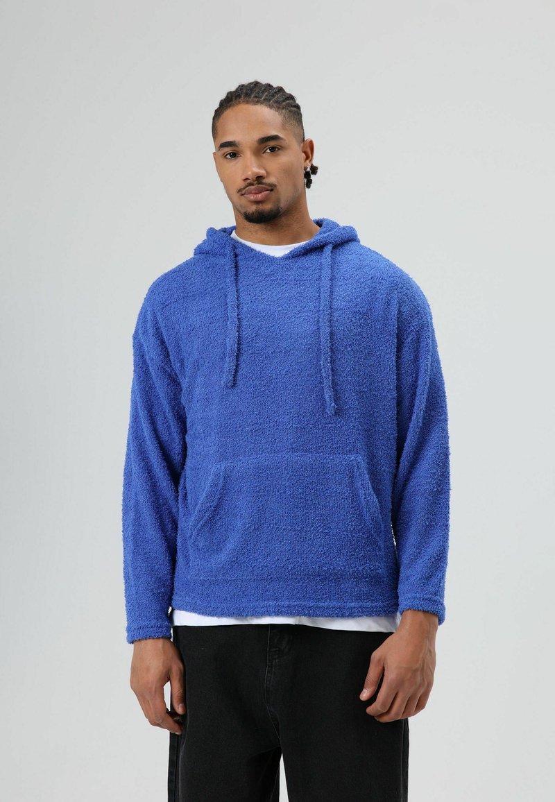 Zachte blauwe hoodie van gestructureerde stof met een voorzak, lange mouwen en een capuchon met trekkoord. Gedragen over een wit shirt.