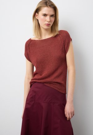 Junge Frau mit glattem blondem Haar, die ein kurzärmeliges rostfarbenes Strickoberteil und einen hoch taillierten dunkelroten Rock trägt, steht vor einer weißen Wand.