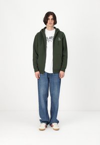 Oliven grønn zip-up hettejakke med doble frontlommer, lagd over en hvit t-skjorte med grafikk, sammen med lyseblå løstsittende jeans og sneakers.