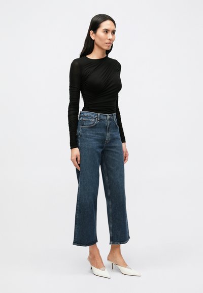 Črna dolgimi rokavi s podrobnostmi nabiranja, kombinirana z modrimi kavbojkami z visokim pasom in širokimi nogami ter belimi mule čevlji s koničastim nosom.