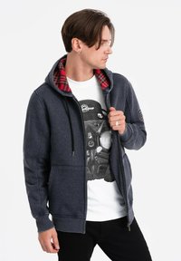 Felpa con zip della marina, foderata in plaid rosso, con tasche frontali e cappuccio regolabile. Indossata sopra una maglietta con grafica e pantaloni neri.