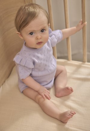 Hellvioletter Strick-Spielanzug mit kurzen Ärmeln und Rüschendetails, Lochmuster und rundem Ausschnitt, sitzend auf einer beigen Stoffoberfläche.