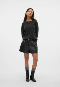 Kvinde iført en mørkegrå sweater, sorte lædershorts med bælte, grå strømper og sorte hælede ankelstøvler, som står foran en ensfarvet baggrund.
