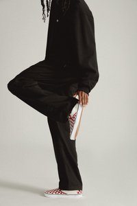 Schwarzes, übergroßes Sweatshirt mit lockerer, schwarzer Hose, die weiße Sneakers mit einem rot-weißen Schachbrettmuster zeigen.