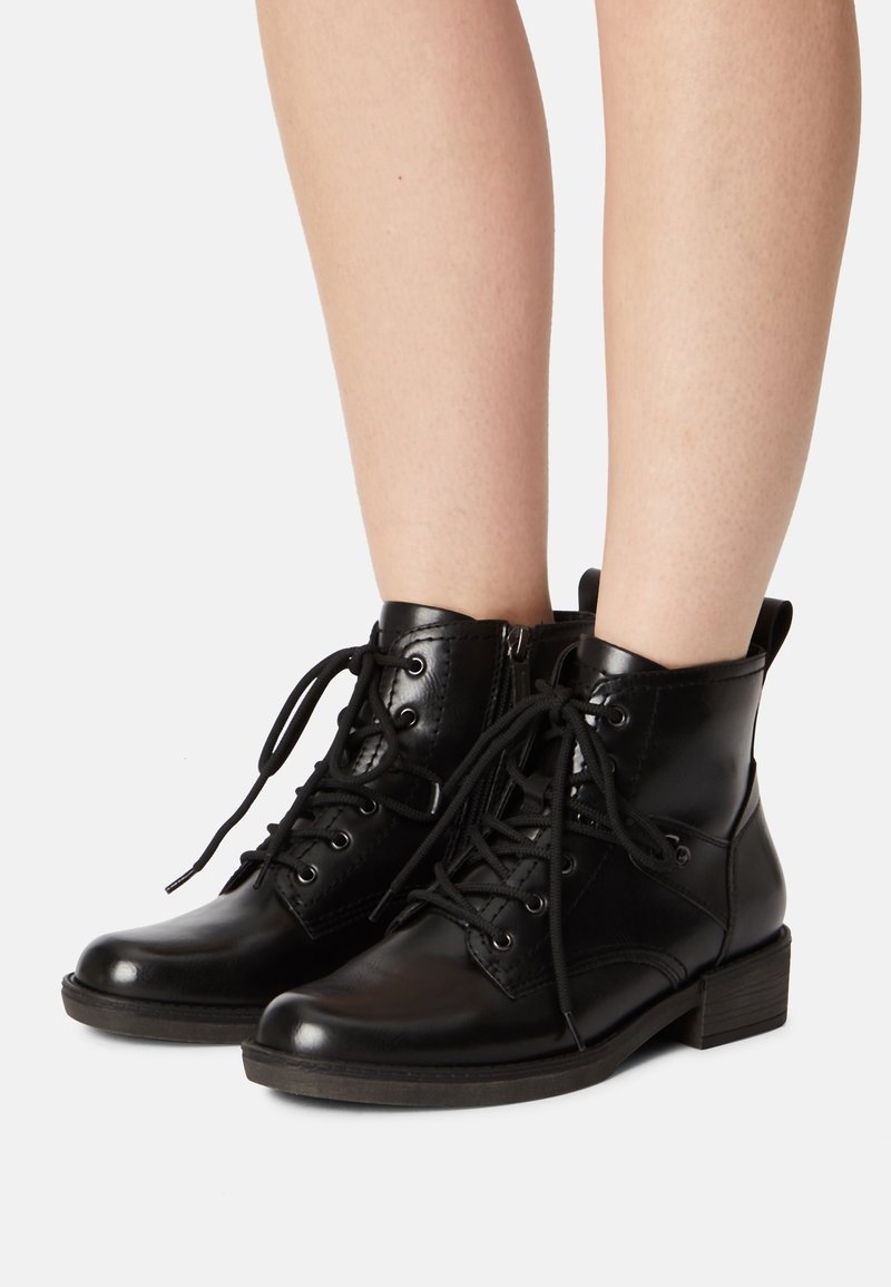 Bottines en cuir noir à lacets jusqu’à la cheville, portées sur des jambes nues, avec fermetures éclair latérales et talons empilés bas sur un fond uni.