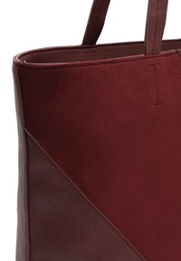 Burgundska usnjena torba z dvema ročkama, ki vključuje teksturiran panel iz zamšene kože in gladke usnjene dodatke, angularno zasnovo in podrobnosti vlakna.