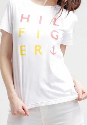 White t-shirt with "HILFIGER" in bold pink and yellow letters and a pink anchor symbol on the right side, worn by a person adjusting the hem.