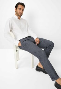 Witte button-up shirt met een borstzak, gecombineerd met donkergrijze broek. Het model zit op een stoel, draagt zwarte schoenen en heeft opgerolde broekspijpen.