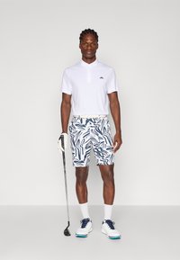 Camisa polo branca com um logótipo preto, calções com padrão em azul e branco, sapatos brancos com detalhes em azul claro, segurando um taco de golfe.