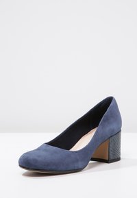 Escarpin en daim bleu marine avec un bout rond et un talon bloc texturé. Le talon présente un accent de motif serpent pour plus de détails.