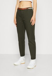 Pantalons vert olive avec une coupe ajustée, ourlets retroussés et ceinture marron. Portés avec des baskets blanches et des chaussettes blanches pour le contraste.