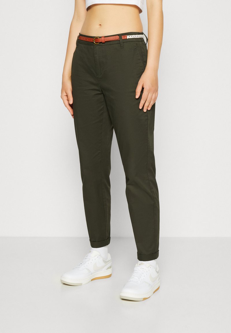 Pantalons vert olive avec une coupe ajustée, ourlets retroussés et ceinture marron. Portés avec des baskets blanches et des chaussettes blanches pour le contraste.