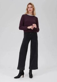 Sweater roxo com um decote de pérolas combinado com calças largas pretas. O modelo usa botins pretos de salto. O fundo é cinza liso.