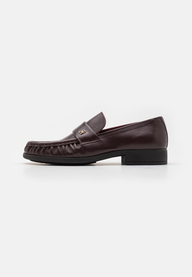 Mocassin en cuir brun foncé avec talon bas, surpiqûres moc toe et petit logo métallique sur la bride.