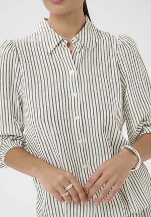 Sieviete valkā baltu un pelēku vertikāli svītrainu pogājamu kreklu ar uzpūstām piedurknēm, sudraba gredzeniem un sudraba aproces rokassprādzi.