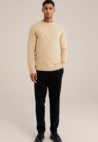 Beige Wollpullover mit Rautenmuster, runder Ausschnitt und gerippten Bündchen, kombiniert mit schwarzen Cordhosen und schwarzen Lederschuhen.
