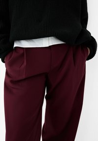 Pantalon bordeaux en tissu lisse, avec des plis devant, une coupe ajustée et des poches latérales, associé à un pull noir à côtes.