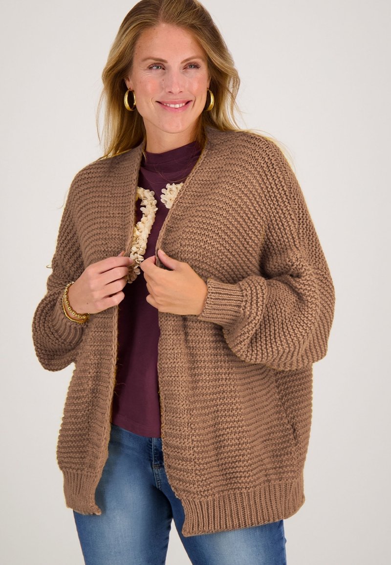 Bruine gebreide cardigan met een textuurpatroon, open voorkant en geribbelde manchetten. Gedragen over een paarse top met een bloemenkettingdetail.