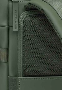 Groene rugzak met een matte afwerking, een gestructureerd mesh zijpaneel en gestikte accenten. Merkenlabel zichtbaar aan de zijkant.