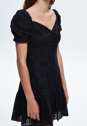 Femme portant une robe en dentelle ajourée noire avec des manches bouffantes, des boutons sur le devant et un ourlet volanté, sur un fond uni.