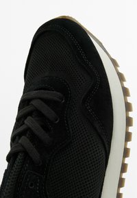Scarpa sportiva nera con tomaia in mesh e dettagli in suede, lacci neri piatti e suola in gomma con un motivo di trazione color cammello.
