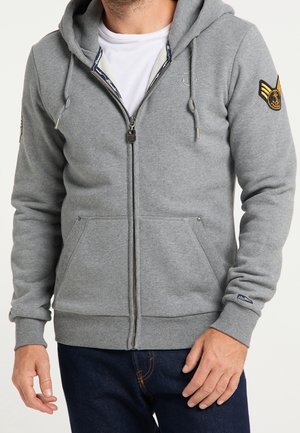 Homme portant un sweat à capuche gris clair zippé avec des poches avant, un patch sur la manche gauche et un t-shirt blanc en dessous, associé à un jean bleu foncé.