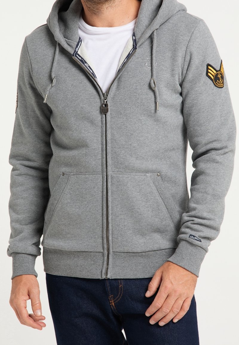 Homme portant un sweat à capuche gris clair zippé avec des poches avant, un patch sur la manche gauche et un t-shirt blanc en dessous, associé à un jean bleu foncé.
