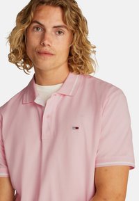 Camisa polo rosa con un collar clásico, tapeta de botones y franjas blancas en contraste en el collar y las mangas. Incluye un parche con el logo.