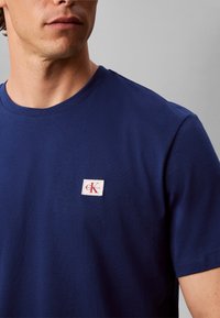 Άνδρας φοράει μπλούζα Calvin Klein σε σκούρο μπλε με μικρό κόκκινο και άσπρο λογότυπο στο στήθος.