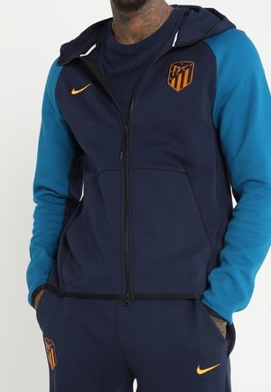 Homme portant un sweat à capuche zippé bleu marine et bleu avec des logos Nike orange et le blason du club de football Atlético Madrid. Mains dans les poches.