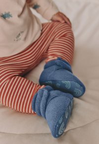 Chaussons en polaire bleue pour bébé avec revers roulé et semelles à motifs, assortis à des leggings rayés rouges et blancs. Texture douce sur un fond clair.