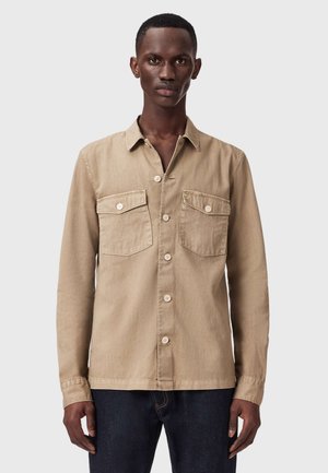 Homme portant une chemise beige à boutons avec deux poches poitrine et un pantalon foncé, debout devant un fond clair uni.