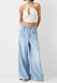 Valkoinen halterneck-crop top, jossa on keskellä aukko, yhdistettynä vaaleansinisiin leveälahkeisiin farkkuihin, joissa on joustava vyötärö ja nyöri.