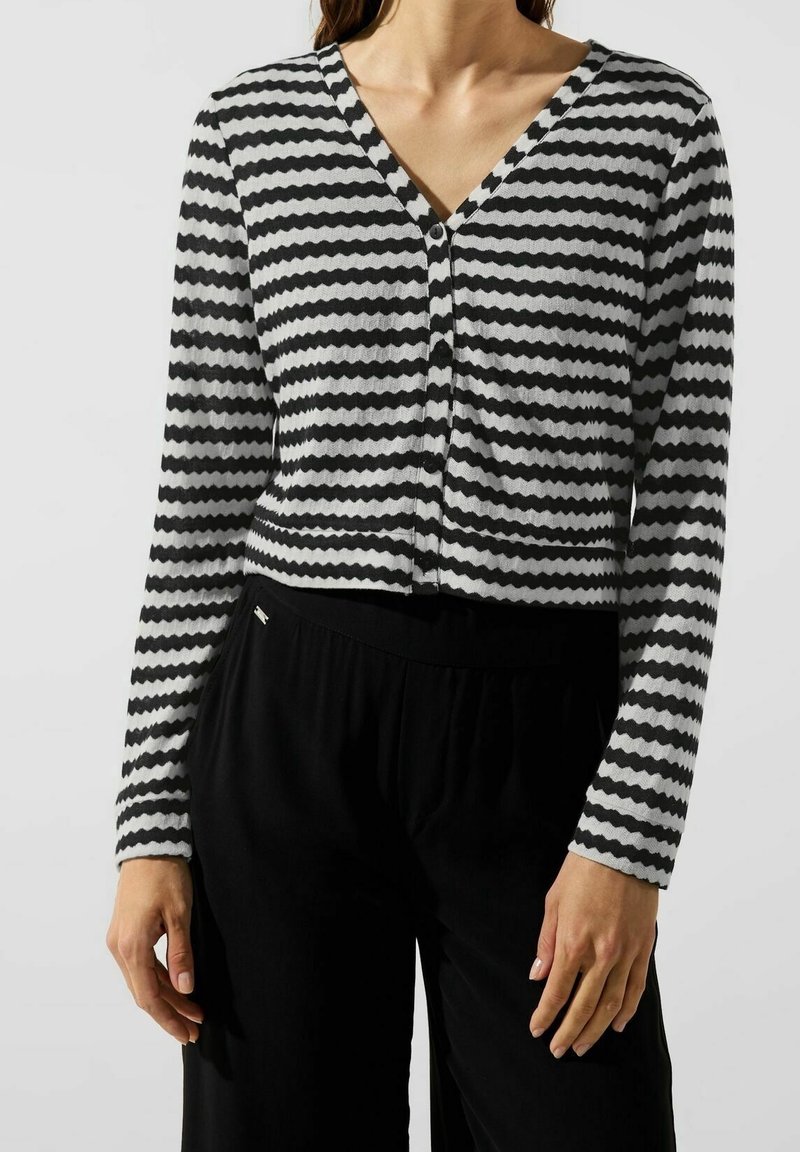 Pruhovaný černobílý cardigan s V-výstřihem, textura s cik-cak vzorem a zapínání na knoflík, v kombinaci s černými, volně střiženými kalhotami.