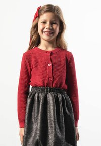 Czerwony dziergany kardigan z zaokrąglonym dekoltem i zapięciem na guziki, w połączeniu z czarną błyszczącą spódnicą z elastycznym paskiem w talii.