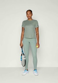 Set di abbigliamento sportivo verde chiaro composto da una t-shirt aderente a maniche corte e leggings a vita alta. Indossa scarpe da ginnastica bianche e tiene in mano una racchetta da tennis.